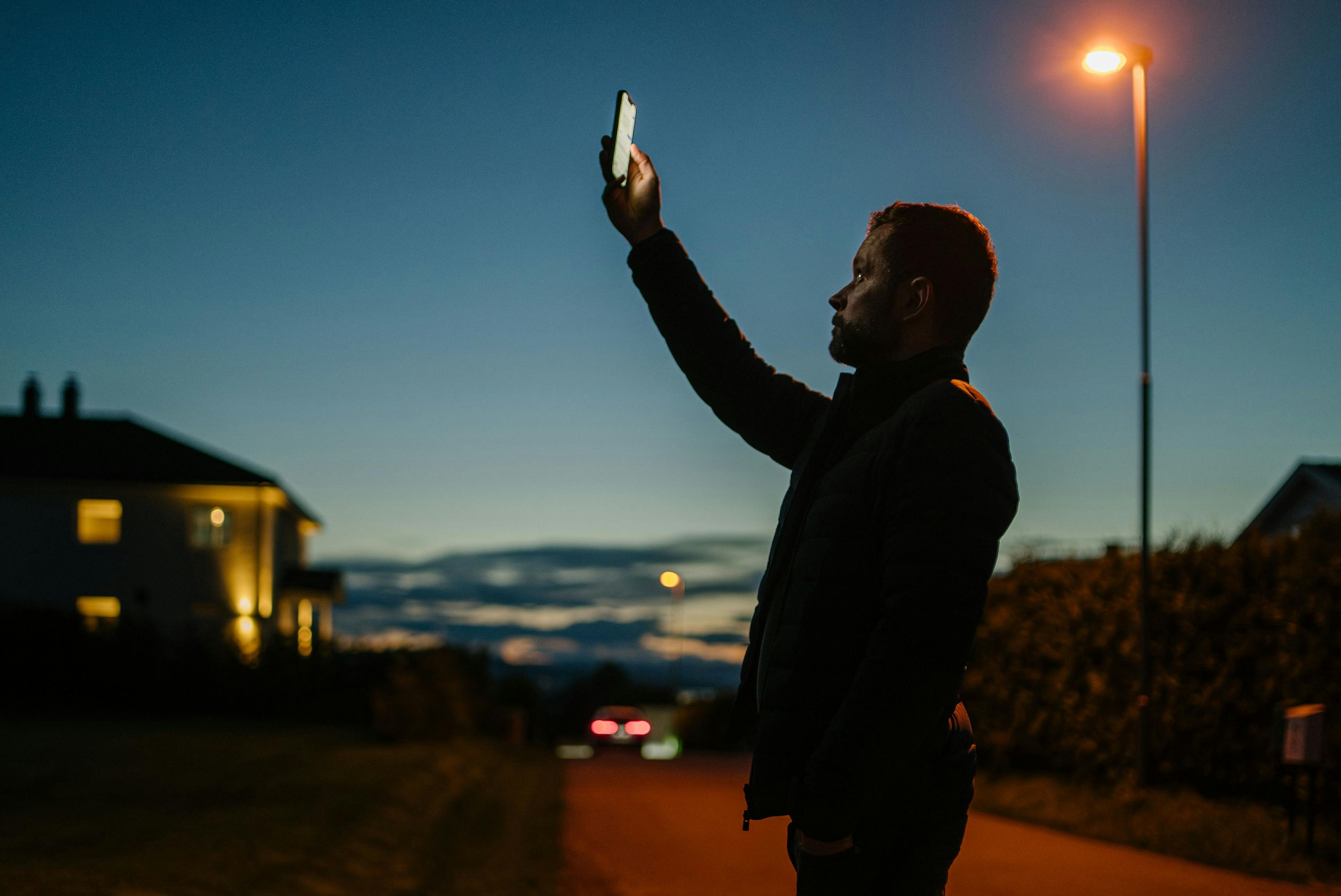 En mann står i et nabolag og holder mobilen høyt for å søke etter dekning.